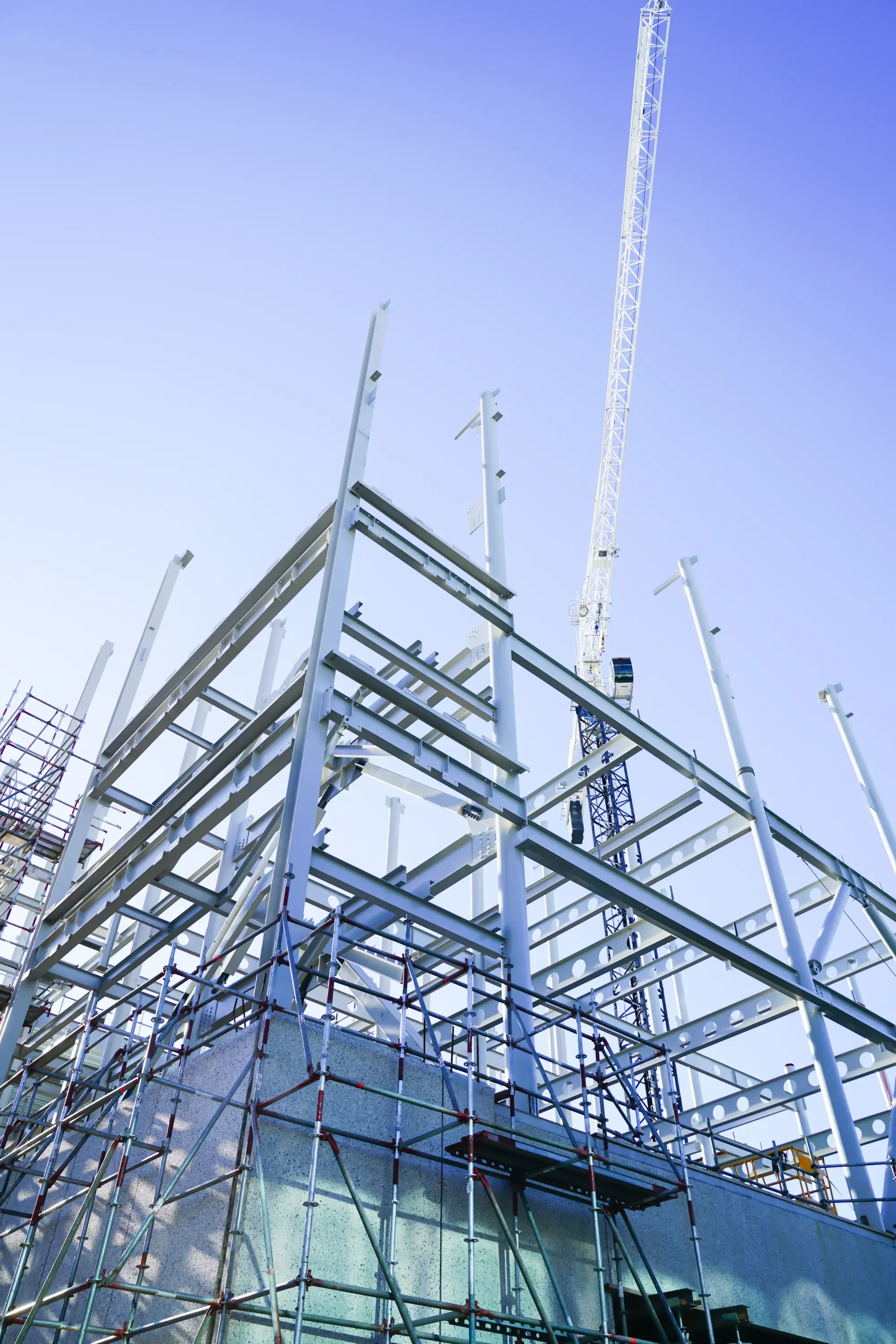Steel-frame commercial building under construction with a tower crane against a clear blue sky